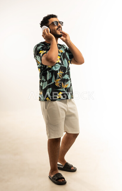 حركات يدين تدل على الرقص والتفاعل مع الموسيقى، بورتريه لشاب عربي خليجي عماني مبتسم يرتدي الزي الكاجوال والنظارة الشمسية يضع سماعات الهيدفون يستمتع بسماع الأغاني المتنوعة، الشعور بالفرح والسعادة، صورة الجسم بالطول الكامل، خلفية بيضاء