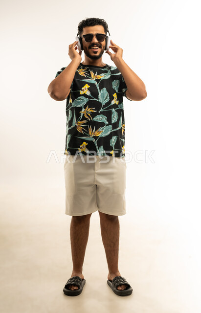 الشعور بالفرح والسعادة، حركات يدين تدل على الرقص والتفاعل مع الموسيقى، بورتريه لشاب عربي خليجي عماني مبتسم يرتدي الزي الكاجوال والنظارة الشمسية يضع سماعات الهيدفون ينظر الى الكاميرا يستمتع بسماع الأغاني المتنوعة، صورة الجسم بالطول الكامل، خلفية بيضاء