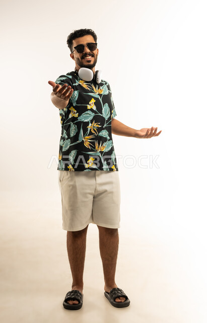 Welcoming and receiving with love and cheerfulness, joy at someone's arrival, portrait of a smiling young Arab Gulf Omani man wearing casual clothes, sunglasses and headphones, opening his hands and looking at the camera with gestures of happiness and joy, full-length body image, white background