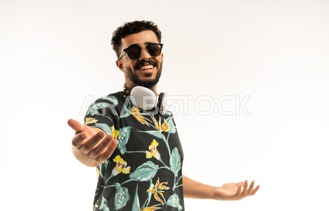 البهجة بقدوم شخص ما, الترحيب والاستقبال بحب وبشاشة، بورتريه مقرب لشاب عربي خليجي عماني مبتسم يرتدي الزي الكاجوال والنظارة الشمسية وسماعة الرأس يفتح كفيه ينظر الى الكاميرا بإيماءات السعادة والفرح، خلفية بيضاء