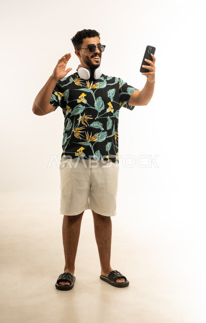 إجراء مكالمة فيديو بتعابير الفرح, دردشة و اتصالات مع الأهل والأصدقاء, بورتريه مقرب لشاب عربي خليجي عماني مبتسم يرتدي الزي الكاجوال والنظارة الشمسية يمسك بيده الهاتف المحمول يرفع يده للاعلى بإيماءات القاء التحية والسلام، صورة الجسم بالطول الكامل، خلفية بيضاء