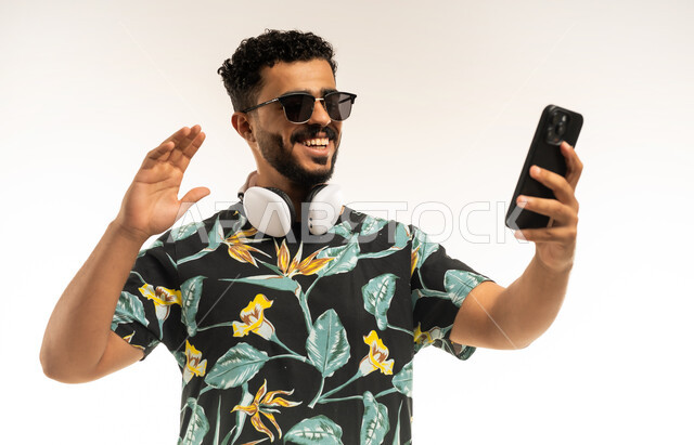 إجراء مكالمة فيديو بتعابير الفرح والسرور, دردشة و اتصالات مع الأهل والأصدقاء, بورتريه مقرب لشاب عربي خليجي عماني مبتسم يرتدي الزي الكاجوال والنظارة الشمسية يمسك الهاتف المحمول يرفع يده للاعلى بإيماءات القاء التحية والسلام، خلفية بيضاء
