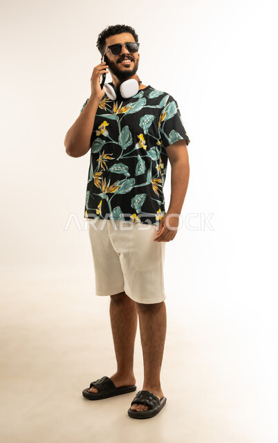 Communication and chatting with family and friends, portrait of a smiling young Arab Gulf Omani man wearing casual clothes and sunglasses holding a mobile phone in his hand making a phone call with gestures of joy and happiness, full body image, white background