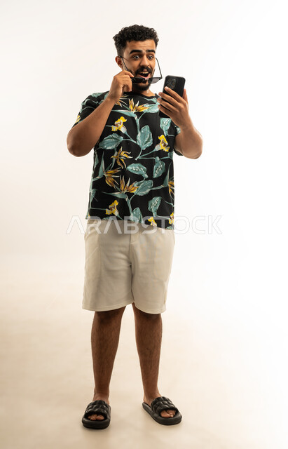 التفاجؤ والصدمة والاستغراب، استخدام التكنولوجيا المتطورة للتسويق الرقمي الالكتروني، بورتريه مقرب لشاب عربي خليجي عماني يرتدي الزي الكاجوال والنظارة الشمسية يمسك بيده الهاتف المحمول ينظر اليه بإيماءات الدهشة والذهول، صورة الجسم بالطول الكامل، خلفية بيضاء