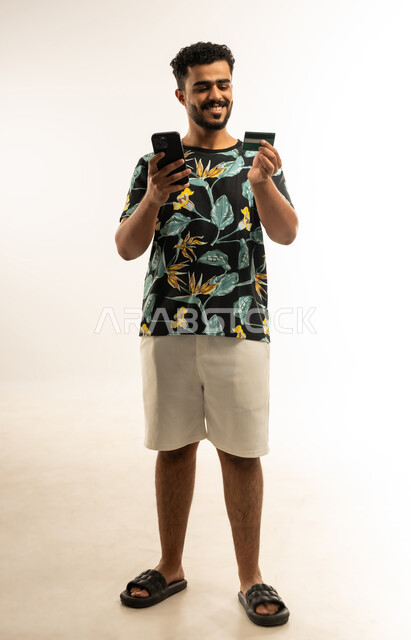 الشراء والدفع من خلال برامج وتطبيقات الجوال، التقنيات الحديثة والمتطورة، بورتريه لشاب عربي خليجي عماني مبتسم يرتدي الزي الكاجوال يمسك بيده الهاتف المحمول ينظر الى بطاقة الإئتمان البنكية بإيماءات الفرح و السرور، صورة الجسم بالطول الكامل، خلفية بيضاء