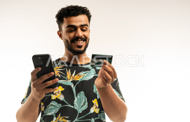 الدفع والشراء من خلال برامج وتطبيقات الجوال، التقنيات الحديثة والمتطورة، بورتريه مقرب لشاب عربي خليجي عماني مبتسم يرتدي الزي الكاجوال يمسك بيده الهاتف المحمول ينظر الى بطاقة الإئتمان البنكية بإيماءات الفرح و السرور، خلفية بيضاء