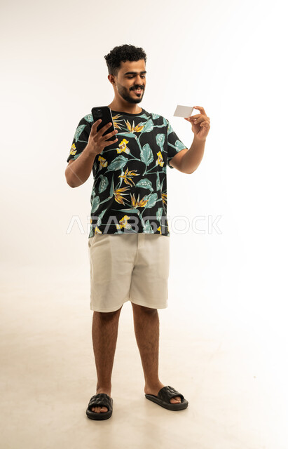 الشراء والدفع من خلال برامج وتطبيقات الجوال، التقنيات الحديثة والمتطورة، بورتريه لشاب عربي خليجي عماني مبتسم يرتدي الزي الكاجوال يمسك بيده الهاتف المحمول ينظر الى بطاقة الإئتمان البنكية بإيماءات الفرح و السرور، صورة الجسم بالطول الكامل، خلفية بيضاء