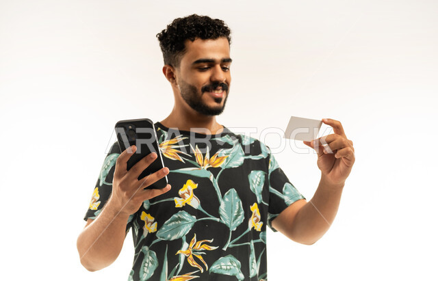 Payment and purchase through mobile programs and applications, modern and advanced technologies, close-up portrait of a smiling young Arab Gulf Omani man wearing casual clothes holding a mobile phone in his hand looking at a bank credit card with gestures of joy and pleasure, white background