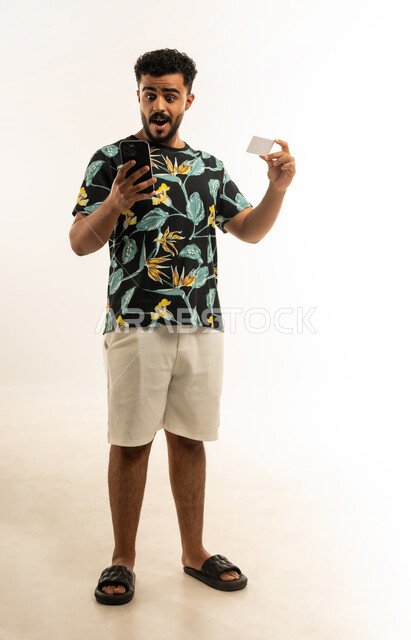 Modern and advanced technologies, purchasing and paying through mobile programs and applications, portrait of a young Arab Gulf Omani man wearing casual clothes holding a mobile phone and a bank credit card, looking at the mobile phone with gestures of surprise and astonishment, full-length body image, white background