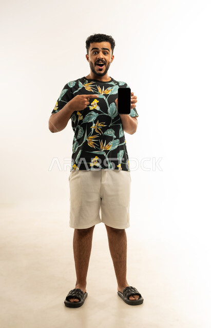 استخدام التكنولوجيا المتطورة للتسويق الرقمي الالكتروني، بورتريه لشاب عربي خليجي عماني يرتدي الزي الكاجوال يمسك بيده الهاتف المحمول بشاشة سوداء فارغة ويشير اليه باصبع السبابة، النظر الى الكاميرا بإيماءات الدهشة والذهول، صورة الجسم بالطول الكامل، خلفية بيضاء