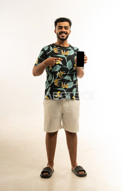 استخدام التكنولوجيا المتطورة للتسويق الرقمي الالكتروني، بورتريه لشاب عربي خليجي عماني مبتسم يرتدي الزي الكاجوال يمسك بيده الهاتف المحمول بشاشة سوداء فارغة ويشير اليه باصبع السبابة، النظر الى الكاميرا بإيماءات السرور، صورة الجسم بالطول الكامل، خلفية بيضاء