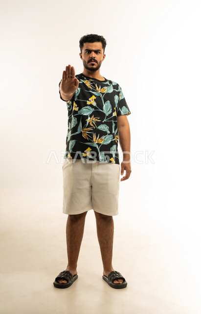 تعابير وإشارات لطلب التوقف و عدم الاقتراب، بورتريه لشاب عربي خليجي عماني يرتدي الزي الكاجوال يرفع يده للأعلى ينظر الى الكاميرا بإيماءات الجدية والحزم، الإمتناع عن فعل شيء ما، صورة الجسم بالطول الكامل، خلفية بيضاء
