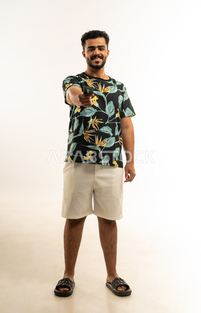 الشراء والدفع عن طريق بطاقات الإئتمان البنكية، إمكانية تسديد الفواتير عبر بطاقة الصراف, بورتريه لشاب عربي خليجي عماني مبتسم يرتدي الزي الكاجوال يمسك بيده البطاقة الالكترونية ينظر الى الكاميرا بإيماءات السرور, صورة الجسم بالطول الكامل، خلفية بيضاء