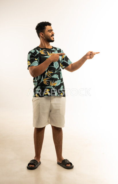 pinpointing, pointing and explaining something, indicating the location of an item, portrait of a smiling young Arab Gulf Omani man in casual wear pointing with index finger at something looking at it with gestures of pleasure, full body image, white background