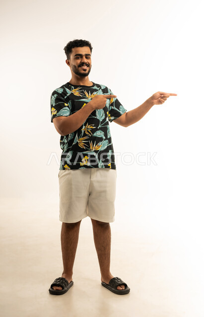 تحديد موقع معين, الإشارة و التوضيح إلى شيء ما, الدلالة على مكان عنصر، بورتريه لشاب عربي خليجي عماني مبتسم يرتدي الزي الكاجوال ينظر الى الكاميرا يشير بإصبع السبابة إلى شيء ما بإيماءات السرور, صورة الجسم بالطول الكامل، خلفية بيضاء