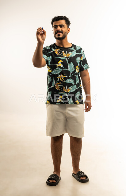 Taking notes, getting ready to write and highlight, portrait of a smiling young Arab Gulf Omani man in casual wear holding a pen writing something with gestures of pleasure, concept of education with prescribed curricula, full body image, white background