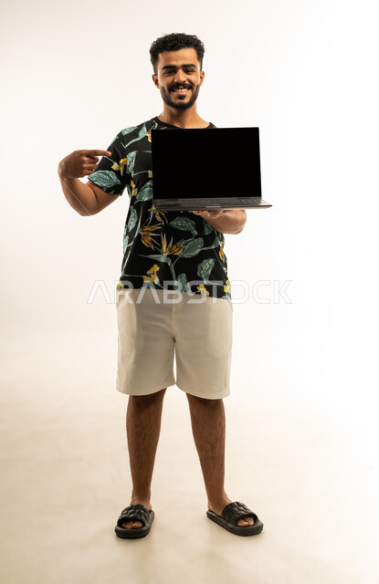 استخدام التقنيات والأجهزة الحديثة، مفهوم إدارة الأعمال عن بعد، بورتريه لشاب عربي خليجي عماني مبتسم يرتدي الزي الكاجوال يحمل بيده جهاز اللابتوب المحمول ينظر الى الكاميرا بإيماءات السرور يشير بإصبع السبابة الى شاشة سواء فارغة, صورة الجسم بالطول الكامل، خلفية بيضاء