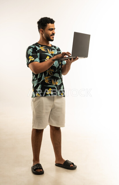 مفهوم إدارة الأعمال عن بعد، اعداد التقارير الخاصة بالعمل، بورتريه لشاب عربي خليجي عماني مبتسم يرتدي الزي الكاجوال يحمل بيده جهاز اللابتوب المحمول، استخدام التقنيات والأجهزة الحديثة لإنجاز المهام، إجراء مكالمات فيديو, صورة الجسم بالطول الكامل، خلفية بيضاء