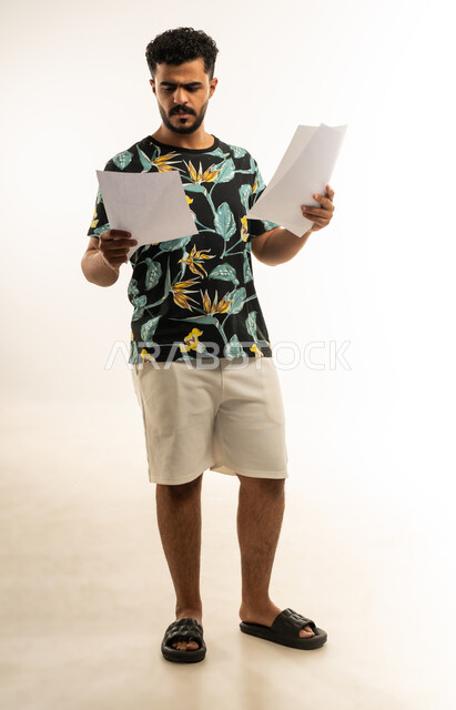 اعداد التقارير الخاصة بالعمل، بورتريه لشاب عربي خليجي عماني مبتسم يرتدي الزي الكاجوال يحمل بيده مجموعة من الاوراق البيضاء، مراجعة ومتابعة الخطط والمشاريع المكتبية بإيماءات التركيز، التدقيق في قراءة الملاحظات، صورة الجسم بالطول الكامل، خلفية بيضاء