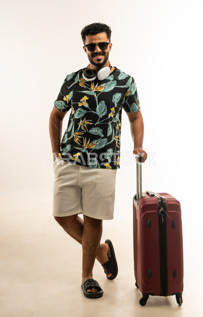 الاستعداد لقضاء رحلة صيفية ممتعة, الذهاب في جولات ترفيهية في إجازة الصيف, تعابير الفرح والسرور، بورتريه لشاب عربي خليجي عماني مبتسم يرتدي الزي الكاجوال والنظارة الشمسية يمسك حقيبة السفر ينظر الى الكاميرا بإيماءات السرور, صورة الجسم بالطول الكامل، خلفية بيضاء