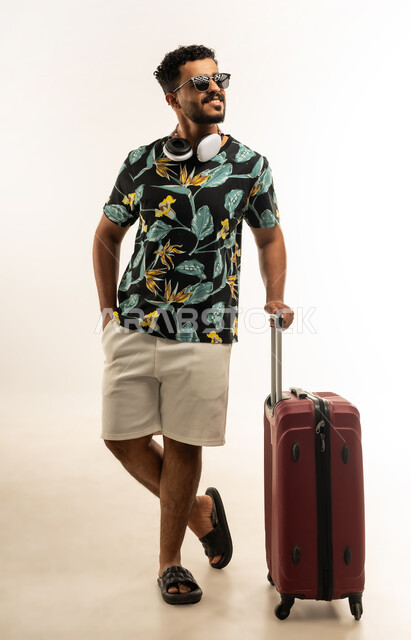 الذهاب في جولات ترفيهية في إجازة الصيف, الاستعداد لقضاء رحلة صيفية ممتعة, تعابير الفرح والسرور، بورتريه لشاب عربي خليجي عماني مبتسم يرتدي الزي الكاجوال والنظارة الشمسية يمسك حقيبة السفر, صورة الجسم بالطول الكامل، خلفية بيضاء