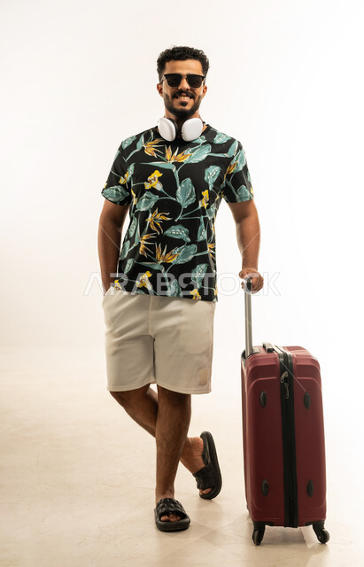 الاستعداد لقضاء رحلة صيفية ممتعة, الذهاب في جولات ترفيهية في إجازة الصيف, تعابير الفرح والسعادة، بورتريه لشاب عربي خليجي عماني مبتسم يرتدي الزي الكاجوال والنظارة الشمسية يمسك حقيبة السفر ينظر الى الكاميرا بإيماءات السرور, صورة الجسم بالطول الكامل، خلفية بيضاء