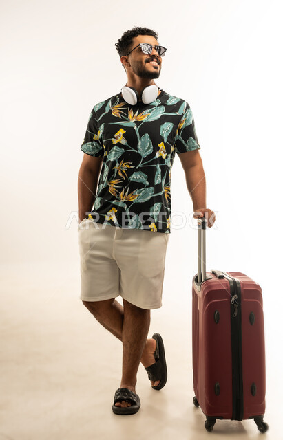الذهاب في جولات ترفيهية في إجازة الصيف, الاستعداد لقضاء رحلة صيفية ممتعة, تعابير الفرح والسرور، بورتريه لشاب عربي خليجي عماني مبتسم يرتدي الزي الكاجوال والنظارة الشمسية يمسك حقيبة السفر, صورة الجسم بالطول الكامل، خلفية بيضاء