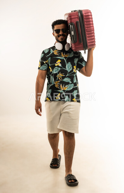الذهاب في جولات ترفيهية في إجازة الصيف, تعابير الفرح والسرور، بورتريه لشاب عربي خليجي عماني مبتسم يرتدي الزي الكاجوال والنظارة الشمسية يحمل حقيبة السفر على كتفه, الاستعداد لقضاء رحلة صيفية ممتعة, صورة الجسم بالطول الكامل، خلفية بيضاء