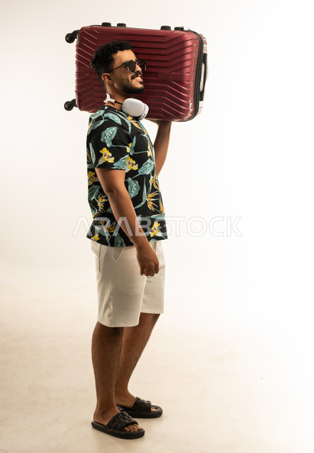 الذهاب في جولات ترفيهية في إجازة الصيف, تعابير الفرح والسرور، بورتريه  لشاب عربي خليجي عماني مبتسم يرتدي الزي الكاجوال والنظارة الشمسية يحمل حقيبة السفر على كتفه, الاستعداد لقضاء رحلة صيفية ممتعة, صورة الجسم بالطول الكامل، خلفية بيضاء