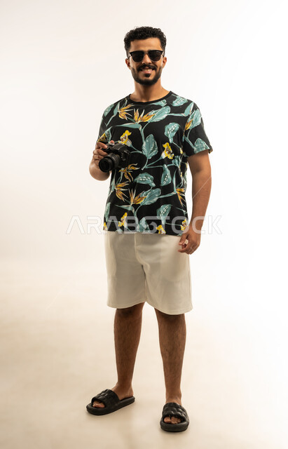 Using high-resolution digital cameras, mastering and professionalizing the photography profession, portrait of a smiling young Arab Gulf Omani man wearing casual clothes and sunglasses looking at the camera with gestures of self-confidence, taking souvenir photos using a photographic camera, full-length body image, white background
