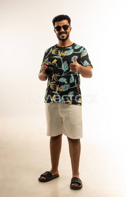 إتقان واحتراف مهنة التصوير، استخدام كاميرات رقمية عالية الدقة، بورتريه  لشاب عربي خليجي عماني مبتسم يرتدي الزي الكاجوال والنظارة الشمسية يرفع اصبع الابهام بعلامة الاعجاب والرضا، التقاط صور تذكارية عبر كاميرا فوتوغرافية، صورة الجسم بالطول الكامل، خلفية بيضاء
