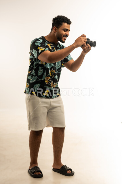 Using high-resolution digital cameras, mastery and professionalism in the photography profession, portrait of a smiling young Arab Gulf Omani man wearing casual clothes taking souvenir photos with a photographic camera, expressions of joy and happiness, full-length body image, white background