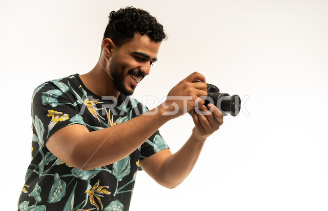 وظائف ومهن سعودية، استخدام كاميرات رقمية عالية الدقة، إتقان واحتراف مهنة التصوير، بورتريه مقرب  لشاب عربي خليجي عماني مبتسم يرتدي الزي الكاجوال يقوم بالتقاط صور تذكارية عبر كاميرا فوتوغرافية، تعابير الفرح والسرور، خلفية بيضاء