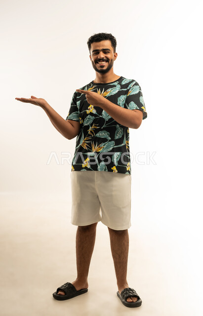 محاولة رفع عنصر ما، مفهوم الاناقة و الاهتمام بالمظهر الخارجي, بورتريه لشاب عربي خليجي عماني مبتسم يرتدي الزي الكاجوال يشير بإصبع السبابة الى كفه يحمل عناصرهولوجرامية ثلاثية الأبعاد ينظر الى الكاميرا بإيماءات السرور، صورة الجسم بالطول الكامل, خلفية بيضاء
