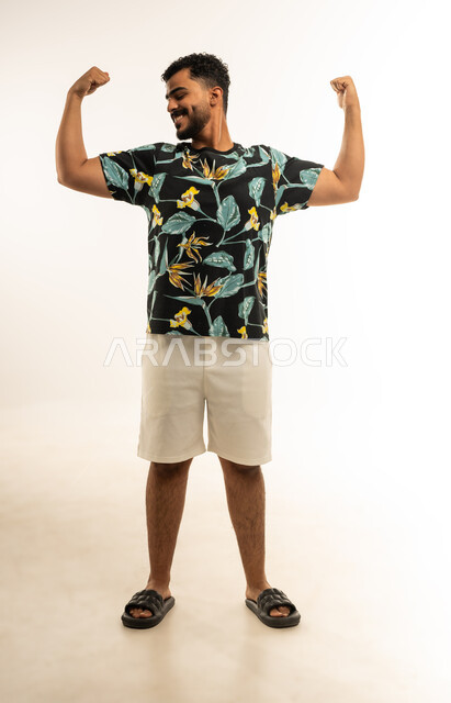 استعراض القوة البدنية، نشاط بدني للمحافظة على صحة الجسم، بناء العضلات وتقويتها، بورتريه لشاب عربي خليجي عماني مبتسم يرتدي الزي الكاجوال يرفع يديه بكفة مقبوضة ينظر الى عضلته، صورة الجسم بالطول الكامل، خلفية بيضاء