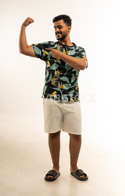 استعراض القوة البدنية، نشاط بدني للمحافظة على صحة الجسم، بناء العضلات وتقويتها، بورتريه لشاب عربي خليجي عماني مبتسم يرتدي الزي الكاجوال يرفع يده بكفة مقبوضة يشير بإصبع السبابة الى عضلته، صورة الجسم بالطول الكامل، خلفية بيضاء