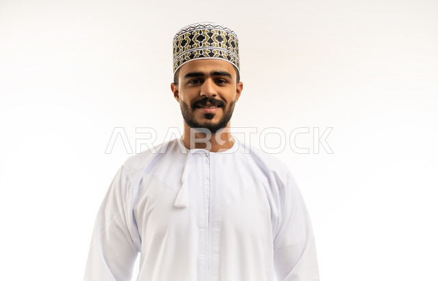 Taking selfies for official documents, interest in elegance and appearance, concept of masculinity and self-confidence, close-up portrait of a smiling young Omani Gulf Arab man wearing a dishdasha and a cloak looking at the camera with gestures of pleasure and happiness, white background