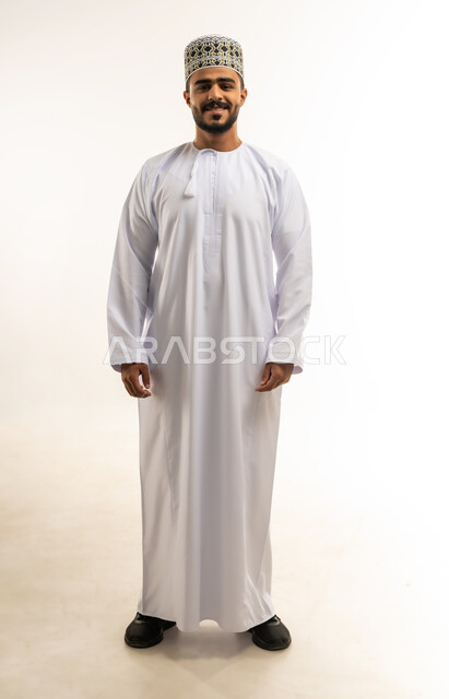 الاهتمام بالأناقة والمظهر الخارجي، التقاط الصور الشخصية للوثائق الرسمية، مفهوم الرجولة والثقة بالنفس، بورتريه مقرب لشاب عربي خليجي عماني مبتسم يرتدي الدشداشة و الكمه ينظر الى الكاميرا بإيماءات السرور والسعادة، صورة الجسم بالطول الكامل، خلفية بيضاء