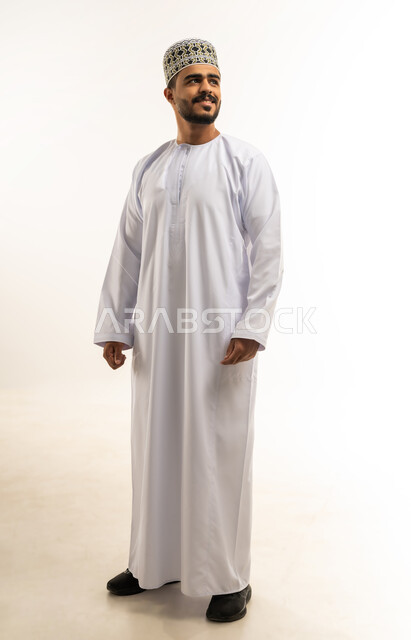 The concept of masculinity and self-confidence, taking personal photos for official documents, portrait of a smiling young Arab Gulf Omani man wearing a dishdasha and a sleeve looking at something with gestures of pleasure and happiness, interest in elegance and external appearance, full-length body image, white background