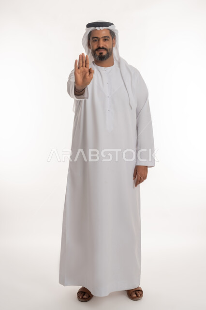 النهي عن فعل شيء ما, الصمت والتوقف عن الكلام, بورتريه لرجل عربي خليجي اماراتي يرتدي الكندورة والغترة الاماراتية يرفع كفه للأعلى بإيماءات الرفض وعدم الرضى والقبول, طلب خفض الصوت والتهديد, صورة الجسم بالطول الكامل, خلفية بيضاء