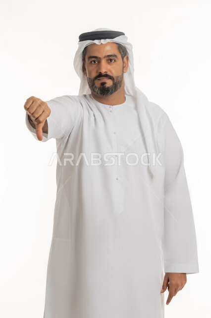 القلق والفشل في تحقيق أمر ما، مفهوم التقييم السلبي والانزعاج، بورتريه لرجل عربي خليجي إماراتي يرتدي الكندورة والغترة ينظر الى الكاميرا يشير بإصبع الابهام الى الاسفل بإيماءات الانزعاج، تعابير الرفض وعدم القبول والرضا، خلفية بيضاء