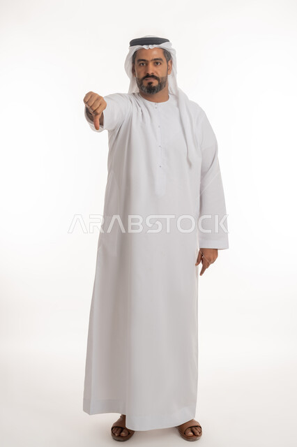 مفهوم التقييم السلبي والانزعاج، القلق والفشل في تحقيق أمر ما، بورتريه لرجل عربي خليجي إماراتي يرتدي الكندورة والغترة ينظر الى الكاميرا يشير بإصبع الابهام الى الاسفل بإيماءات الانزعاج، تعابير الرفض وعدم القبول والرضا، صورة الجسم بالطول الكامل، خلفية بيضاء