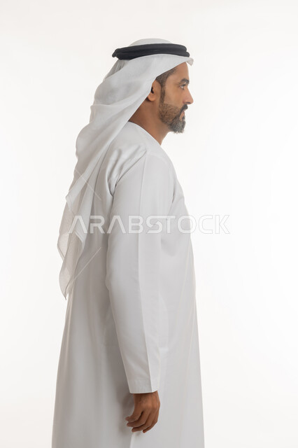 الوقوف باستقامه بتعابير الثقه بالنفس، مفهوم الاهتمام بالمظهر الخارجي والاناقه، بورتريه من الجانب لرجل عربي خليجي اماراتي يرتدي الكندوره والغتره البيضاء يقف بايماءات الرجولة، خلفية بيضاء