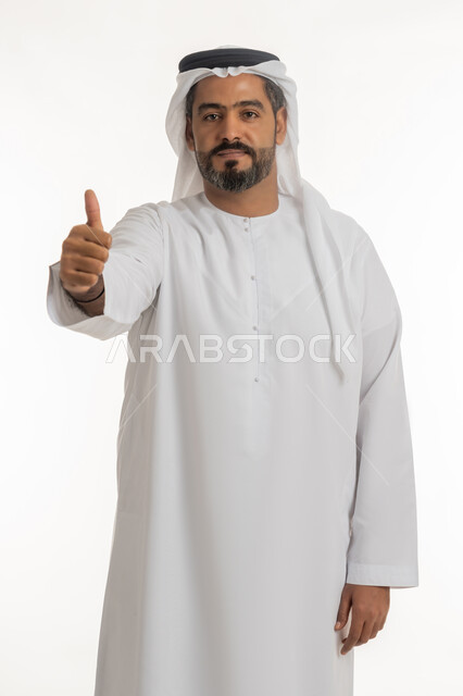 الاهتمام بالأناقة والمظهر الخارجي، ايماءات القبول والفرح والسرور، بورتريه لرجل عربي خليجي إماراتي يرتدي الكندورة والغترة ينظر الى الكاميرا يرفع إصبع السبابة للاعلى بتعابير الإعجاب والرضا، خلفية بيضاء