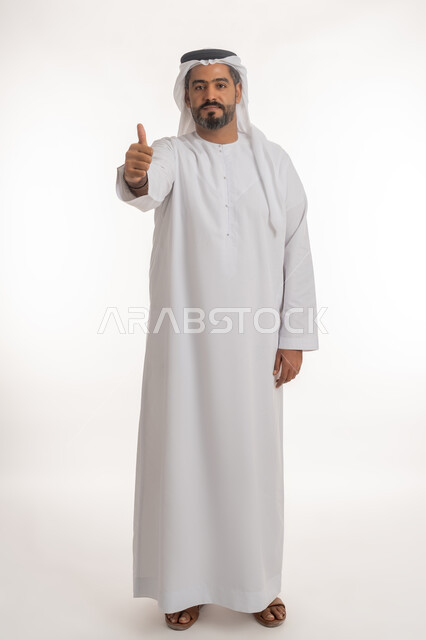 ايماءات القبول والفرح والسرور، الاهتمام بالأناقة والمظهر الخارجي، بورتريه لرجل عربي خليجي إماراتي يرتدي الكندورة والغترة ينظر الى الكاميرا يرفع إصبع السبابة للاعلى بتعابير الإعجاب والرضا، الجسم بالطول الكامل, خلفية بيضاء