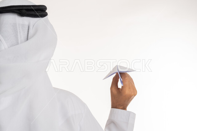 الوصول الى الهدف والتحقيق, التطلع للمستقبل و تطوير الأفكار, بورتريه مقرب من الخلف لرجل عربي خليجي اماراتي مبتسم يرتدي الكندورة والغترة الاماراتية يحمل طائرة مصنوعة من الورق, خلفية بيضاء