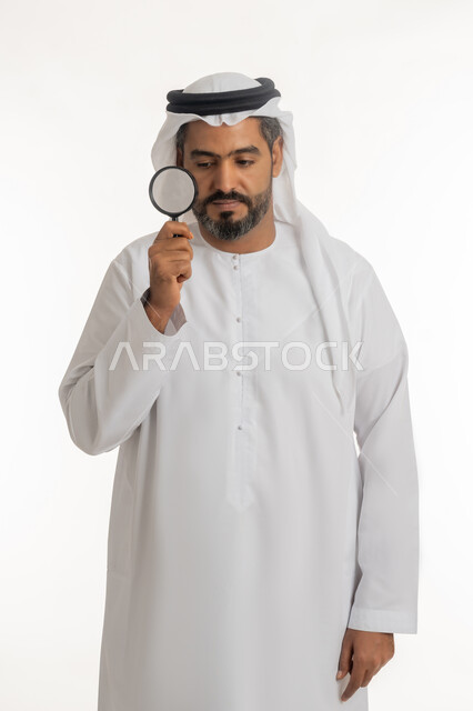 توضيح الصورة والرؤية عن قرب، مفهوم الفحص والاستكشاف، بورتريه لرجل عربي خليجي اماراتي يرتدي الكندورة والغترة الاماراتية ينظر من العدسة مكبرة بإيماءات التركيز والتمعن, خلفية بيضاء