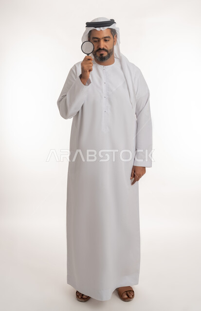 مفهوم الفحص والاستكشاف، توضيح الصورة والرؤية عن قرب، بورتريه لرجل عربي خليجي اماراتي يرتدي الكندورة والغترة الاماراتية ينظر من العدسة مكبرة بإيماءات التركيز والتمعن، الجسم بالطول الكامل، خلفية بيضاء