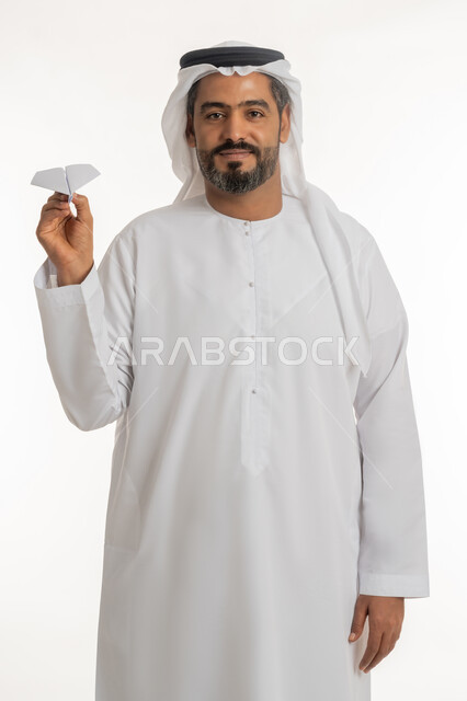 النظر الى الكاميرا بايماءات الفرح والسرور، التحقيق والوصول الى الهدف, تطوير الأفكار و التطلع للمستقبل, بورتريه لرجل عربي خليجي اماراتي مبتسم يرتدي الكندورة والغترة الاماراتية يحمل طائرة مصنوعة من الورق, خلفية بيضاء