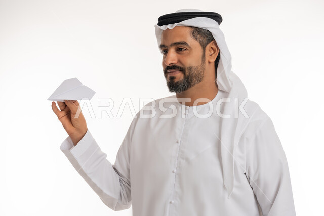 تطوير الأفكار و التطلع للمستقبل, مخطط طائرة أوريغامي بأجنحة، التحقيق والوصول الى الهدف, ايماءات الفرح والسرور، بورتريه مقرب لرجل عربي خليجي اماراتي مبتسم يرتدي الكندورة والغترة الاماراتية يحمل طائرة مصنوعة من الورق, خلفية بيضاء